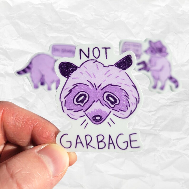 以下が含まれることがあります： 白い背景に「NOT GARBAGE」という文字が書かれた、紫色のアライグマのステッカーです。