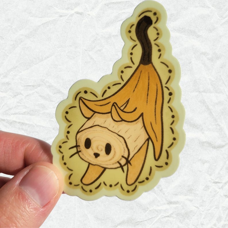 Puede incluir: Una pegatina de un gato de dibujos animados que lleva una c&aacute;scara de pl&aacute;tano como sombrero. El gato es marr&oacute;n y blanco y tiene una expresi&oacute;n linda. La c&aacute;scara de pl&aacute;tano es amarilla y tiene un tallo marr&oacute;n. La pegatina est&aacute; sobre un fondo blanco.