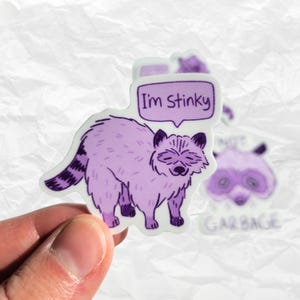以下が含まれることがあります： 紫色のアライグマのステッカーで、吹き出しの中に「I'm Stinky」という文字が書かれています。アライグマは四つん這いで立っており、かわいい表情をしています。