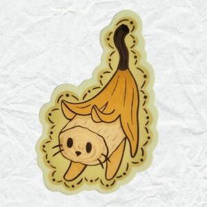Puede incluir: Una c&aacute;scara de pl&aacute;tano amarilla se utiliza como sombrero para una figurita de gato de madera. El gato tiene una cara marr&oacute;n y blanca con ojos negros y bigotes. La figurita est&aacute; rodeada por un borde decorativo amarillo y marr&oacute;n.