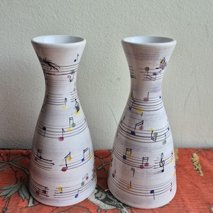 Może przedstawiać: Dwa białe wazony ceramiczne z motywami muzycznymi. Wazony zdobione są czarnymi nutami i kolorowymi akcentami. Mają rozkloszowaną górę i smukły korpus. Tło to czerwona tkanina we wzory.