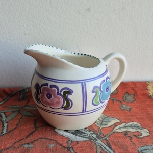 May include: A white ceramic creamer with a floral design in shades of purple, blue, and green. The creamer has a rounded body and a handle.