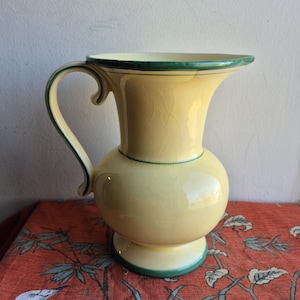 May include: A yellow ceramic pitcher with a rounded body and a flared top. The pitcher has a curved handle and is trimmed with green paint around the rim, base, and handle. The pitcher is sitting on a red patterned surface.