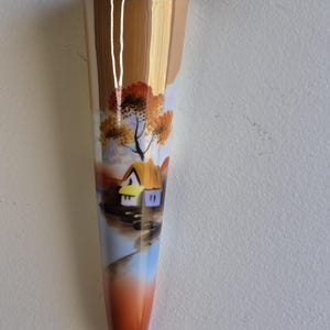 May include: A slender, hand-painted vase with a landscape scene. The vase features a brown and gold exterior with a painted scene of a house, trees, and water in shades of orange, blue, and brown. The vase is approximately 8 inches tall.
