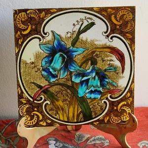 Può includere: Una piastrella di ceramica vintage con un motivo floreale. La piastrella presenta un fiore blu e verde con accenti gialli e marroni. La piastrella è incorniciata da un bordo dorato e marrone.