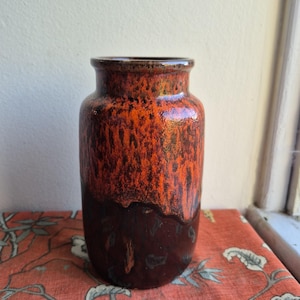 May include: A cylindrical ceramic vase featuring a glossy glaze with a vibrant orange upper section that transitions into a deep, dark brown base. The surface displays a textured, dripped effect where the two colors meet, creating a distinct contrast.