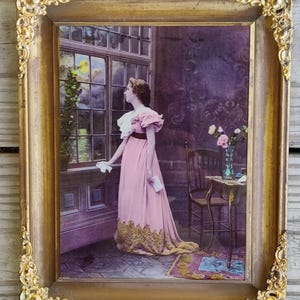 May include: A framed vintage print of a woman in a pink dress standing by a window. The woman is looking out the window and holding a letter. The print is in a gold frame with ornate details.