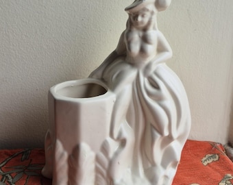 Puede incluir: Macetero de cerámica blanca con forma de mujer con vestido y sombrero. El macetero tiene una abertura hexagonal para plantar.