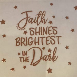 May include: A light-colored background is adorned with numerous bronze-colored stars of varying sizes. The phrase "Faith SHINES BRIGHTEST IN THE Dark" is written in a decorative bronze font, adding a touch of inspiration.