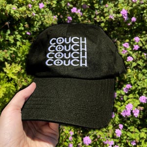 Könnte beinhalten: Schwarze Baseballkappe mit dem Wort "COUCH" in Weiß gestickt, viermal wiederholt. Jedes Wort hat ein Fadenkreuz-Symbol über dem "O". Die Kappe wird vor einem Hintergrund aus grünen Blättern und lila Blumen gehalten.