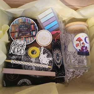 Op de afbeelding: Een geschenkdoos gevuld met briefpapier en accessoires. Inclusief een glazen pot met een paddenstoel- en maanontwerp, stickers, een bladwijzer, een pen, een scrunchie en plaknotities. De doos is gevoerd met geel vloeipapier.