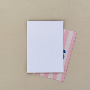 May include: A blank white rectangular card rests atop a smaller card with pink and white stripes. The corner of the striped card has blue text. The background is a solid, light beige color.