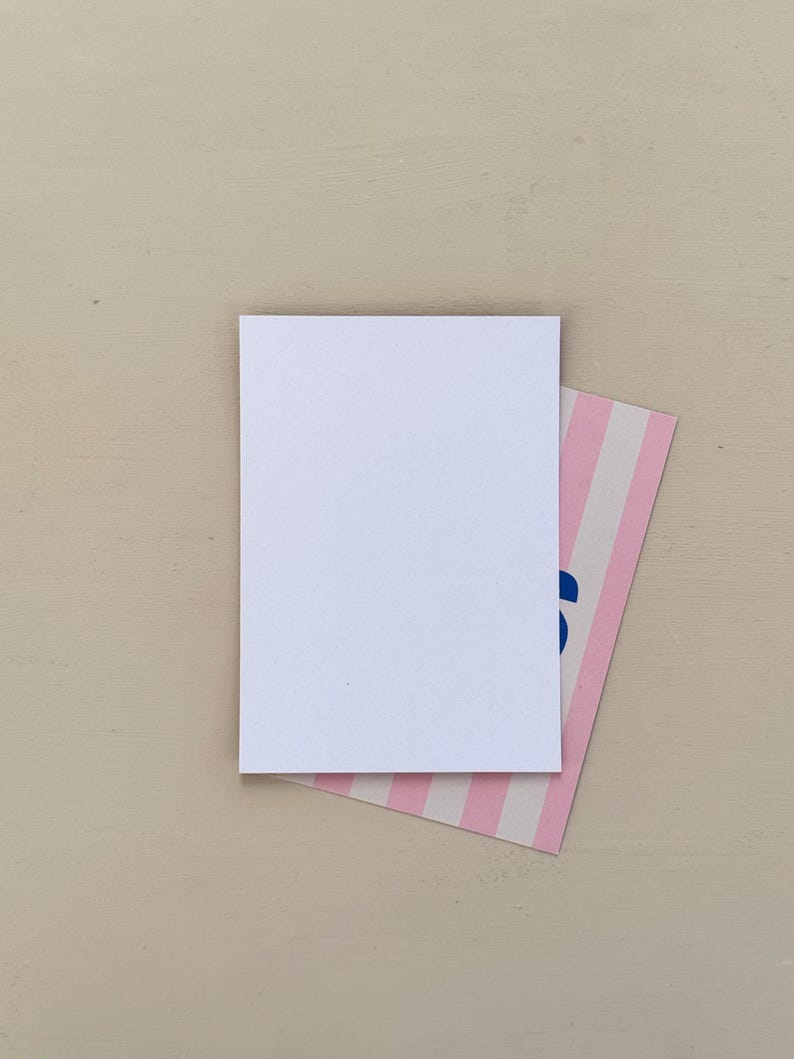 May include: A blank white rectangular card rests atop a smaller card with pink and white stripes. The corner of the striped card has blue text. The background is a solid, light beige color.