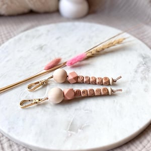 May include: Two wooden bead keychains with the name "UROMA" spelled out in wooden letter beads. The keychains have gold-colored clasps and are accented with pink and peach-colored beads. The keychains are displayed on a white marble surface with dried flowers.