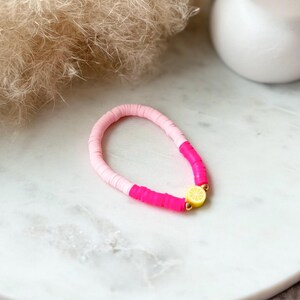 May include: A handmade bracelet featuring a mix of pink and hot pink disc beads. A yellow lemon charm with gold-colored accents adds a pop of color. The bracelet is displayed on a white marble surface.