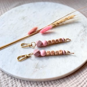May include: Two keychains with the word "MAMA" spelled out in wooden letter beads. The keychains feature pink and mauve-colored beads and gold-colored clasps. The keychains are displayed on a white marble surface with dried flowers.