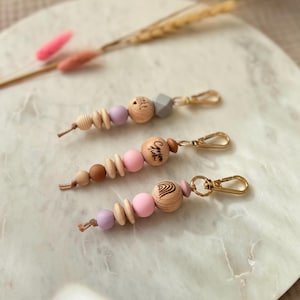 May include: Three handmade keychains with wooden beads in natural, pink, and lavender hues. Each keychain features a gold-colored clasp and a variety of bead shapes, including round and geometric. One keychain has the word "Chloe" engraved on it.