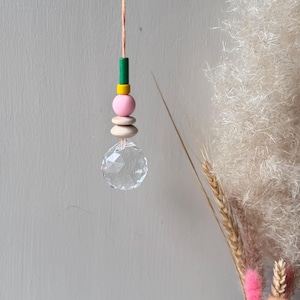 Könnte beinhalten: Ein hängender Kristall-Sonnenfänger mit einer Schnur aus bunten Holzperlen. Die Perlen sind grün, gelb, rosa und in Naturholztönen gehalten. Am unteren Ende hängt eine klare, facettierte Kristallkugel. Der Hintergrund umfasst Trockenblumen und eine hellgraue Wand.