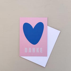 May include: A pink greeting card with a blue heart design and the word "DANKE" in white. A white envelope is partially visible behind the card. The card is on a light beige surface.