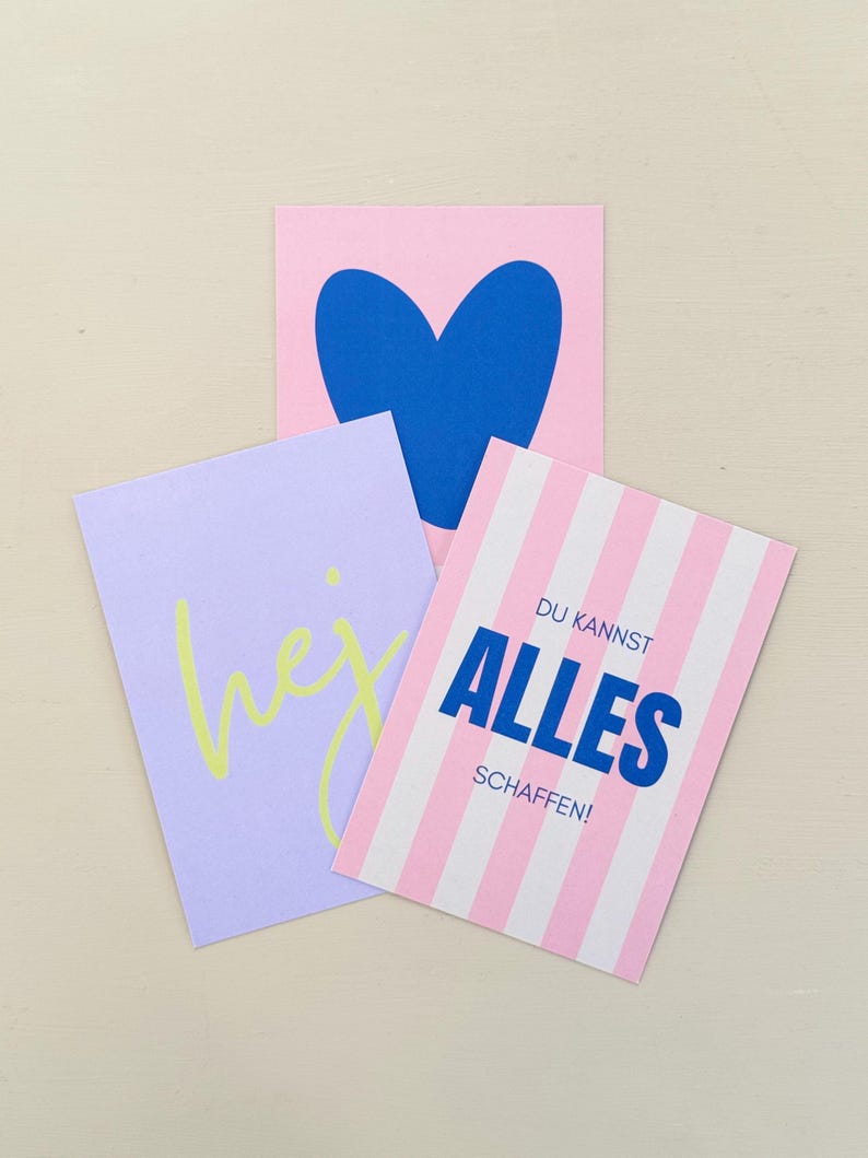 May include: Three colorful cards with graphic designs. One pink card features a blue heart. A lavender card has the word "hej" in yellow. A striped pink and white card has the text "DU KANNST ALLES SCHAFFEN!" in blue.