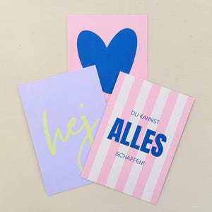 May include: Three colorful cards with graphic designs. One pink card features a blue heart. A lavender card has the word "hej" in yellow. A striped pink and white card has the German text "DU KANNST ALLES SCHAFFEN!"