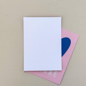 May include: A white rectangular card rests atop a pink card with a blue heart design. The pink card has the word "MAKE" printed in white. The background is a light beige color.