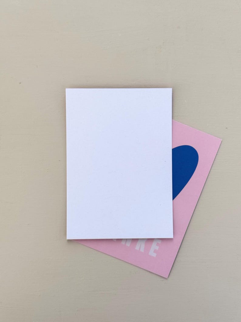 May include: A white rectangular card rests atop a pink card with a blue heart design. The pink card has the word "MAKE" printed in white. The background is a light beige color.