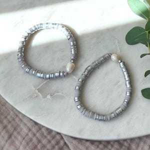 May include: Two gray and white beaded bracelets with a pearl accent. The bracelets are on a white marble surface.