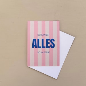 May include: A greeting card with pink and white vertical stripes. The card has the words "DU KANNST ALLES SCHAFFEN!" in blue. A white envelope is partially visible behind the card.