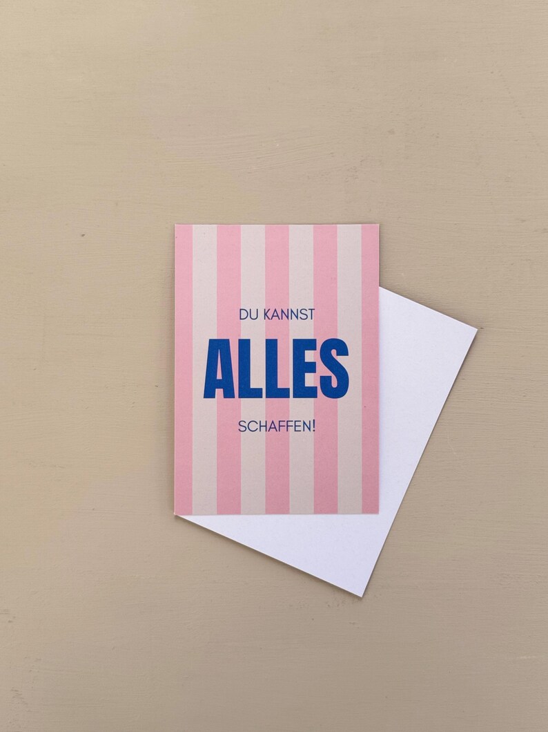 May include: A greeting card with pink and white vertical stripes. The card has the words "DU KANNST ALLES SCHAFFEN!" in blue. A white envelope is partially visible behind the card.