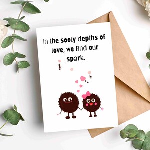 May include: A greeting card with the text "In the sooty depths of love, we find our spark." features two cartoon characters holding hands. The card is on a brown envelope, surrounded by white flowers and green leaves.