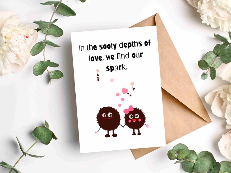 May include: A greeting card with the text "In the sooty depths of love, we find our spark." features two cartoon characters holding hands. The card is on a brown envelope, surrounded by white flowers and green leaves.