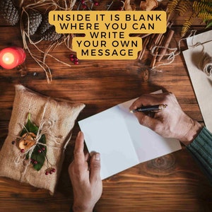 May include: A person writing in a blank greeting card on a wooden surface. The card is next to a wrapped gift, a lit candle, pine cones, and other decorative items. Text on the image reads: "INSIDE IT IS BLANK WHERE YOU CAN WRITE YOUR OWN MESSAGE."