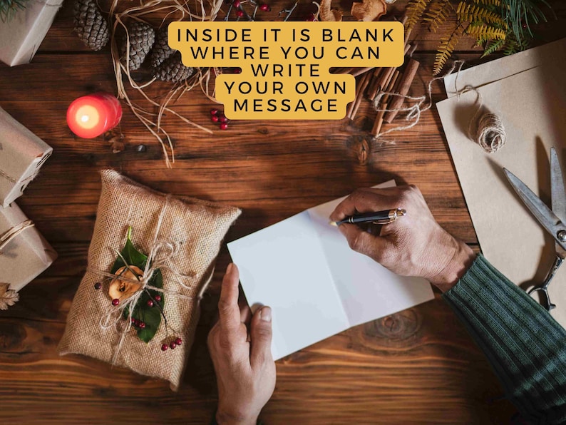 May include: A person writing in a blank greeting card on a wooden surface. The card is next to a wrapped gift, a lit candle, pine cones, and other decorative items. Text on the image reads: "INSIDE IT IS BLANK WHERE YOU CAN WRITE YOUR OWN MESSAGE."