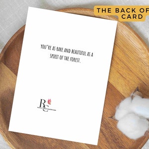 May include: A white greeting card with the text "YOU'RE AS RARE AND BEAUTIFUL AS A SPIRIT OF THE FOREST." is displayed on a wooden plate. The words "THE BACK OF THE CARD" are at the top. A cotton flower is on the right.