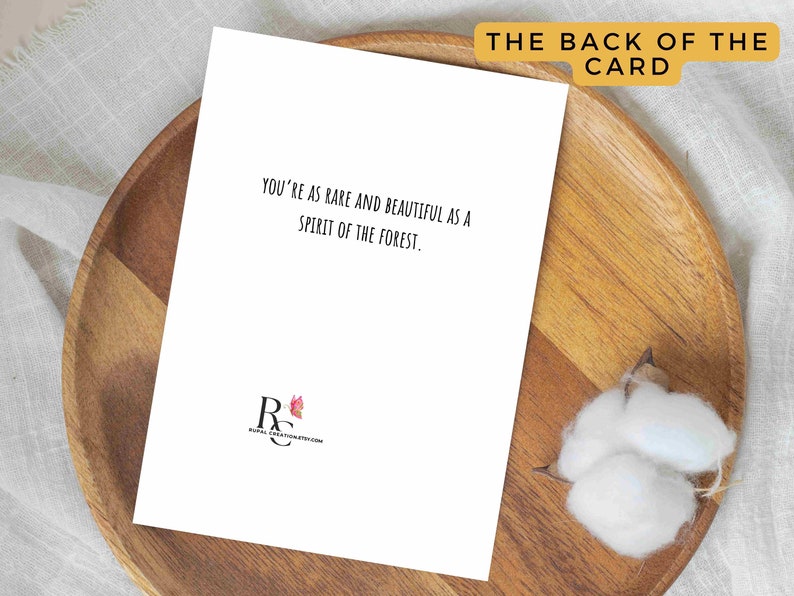 May include: A white greeting card with the text "YOU'RE AS RARE AND BEAUTIFUL AS A SPIRIT OF THE FOREST." is displayed on a wooden plate. The words "THE BACK OF THE CARD" are at the top. A cotton flower is on the right.