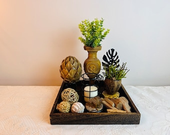 May include: A wooden tray with a variety of decorative items, including a candle, a small potted plant, a wooden vase with a green plant, and a wooden artichoke. The tray is on a white tablecloth.