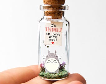 May include: A small glass bottle with a cork stopper. Inside, a Totoro character illustration and the text "I'm TOTOROLY in love with you!" are visible. The bottle contains small flowers and greenery.