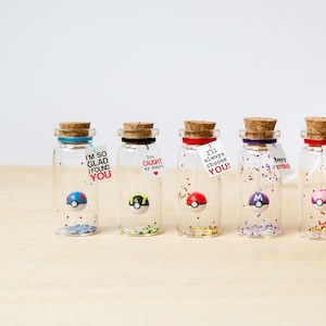 May include: Five small clear glass bottles with cork stoppers, each containing a miniature spherical toy and colourful glitter. Each bottle features a small white tag with printed text including phrases like I'm so glad I found you and I love you.