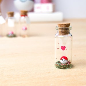 May include: A small glass jar with a cork lid contains a red and white Pokeball with green glitter. The jar has red hearts and red dots painted on the side.