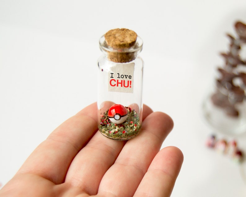 May include: A small glass jar with a cork lid filled with green glitter and a red and white Pokeball. A small white note with the text "I love CHU!" is attached to the jar.