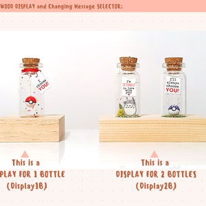 May include: Two wooden display stands for small glass bottles. The stand on the left holds one bottle with a red and white Pokeball inside. The stand on the right holds two bottles, one with a Totoro character inside and the other with a Pokeball inside. The text on the stands reads "This is a DISPLAY FOR 1 BOTTLE (Display16)" and "This is a DISPLAY FOR 2 BOTTLES (Display26)".