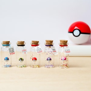 May include: Five small clear glass bottles with cork stoppers, each containing a miniature spherical toy and colorful glitter. Each bottle features a small white tag with printed text including phrases like I'm so glad I found you and I love you.