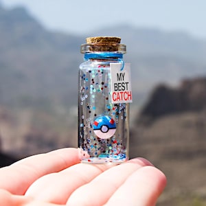 May include: A small clear glass bottle with a cork stopper containing blue, red, and silver glitter and a miniature blue and white ball-shaped charm. A white rectangular tag tied with blue string features the black and red text MY BEST CATCH.