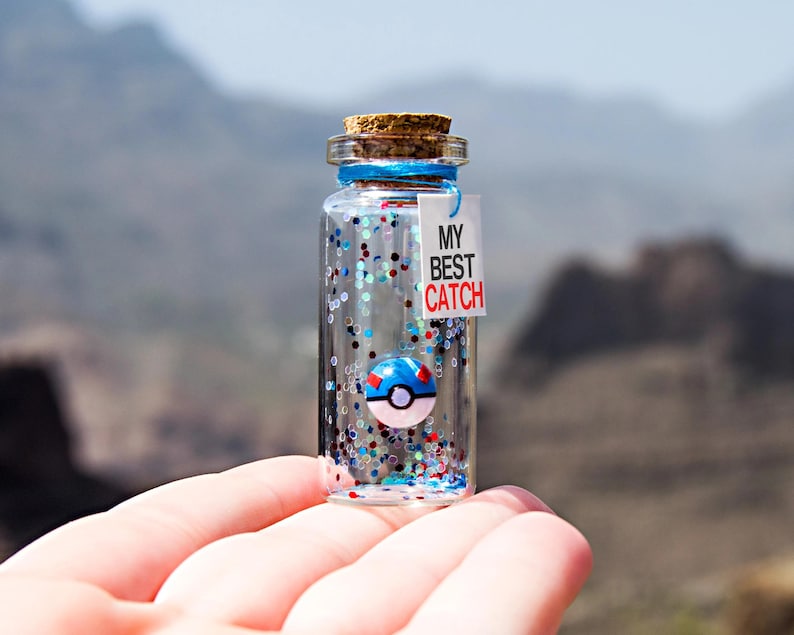 May include: A small clear glass bottle with a cork stopper containing blue, red, and silver glitter and a miniature blue and white ball-shaped charm. A white rectangular tag tied with blue string features the black and red text MY BEST CATCH.