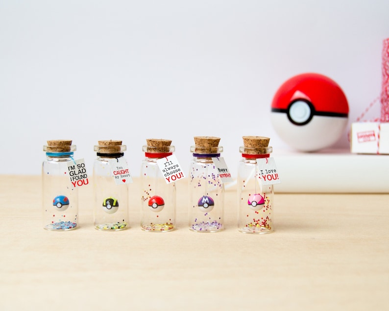 May include: Six small glass bottles with cork tops, each containing a small Pokeball charm and colorful glitter. The bottles have labels with different messages, including "I'm so glad I found you", "I'll always choose you", and "I love you".