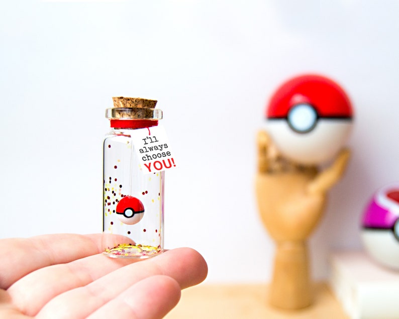 May include: A small clear glass bottle with a cork stopper containing a miniature red and white ball, gold and red glitter, and a white paper tag tied with a red string that features the black and red text I'll always choose YOU!