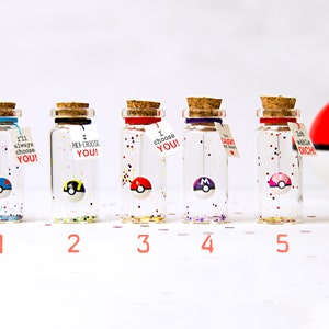 May include: Five clear glass bottles with cork stoppers, each containing a small spherical ball resembling a Pok&eacute; Ball and colorful glitter. Each bottle features a small white tag with text including I'll always choose YOU, I PKA-CHOOSE YOU, I choose YOU, You caught my heart, and Ich w&auml;hle DICH.