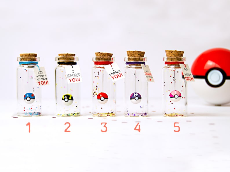 May include: Five clear glass bottles with cork stoppers, each containing a small spherical ball resembling a Pok&eacute; Ball and colorful glitter. Each bottle features a small white tag with text including I'll always choose YOU, I PKA-CHOOSE YOU, I choose YOU, You caught my heart, and Ich w&auml;hle DICH.