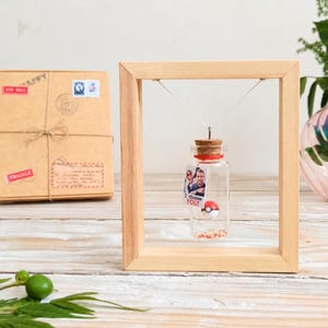 May include: A small glass bottle with a cork stopper hangs inside a light-colored wooden frame. Inside the bottle is a tiny red and white ball and a small rectangular tag featuring a photograph of two people and the text I choose YOU!